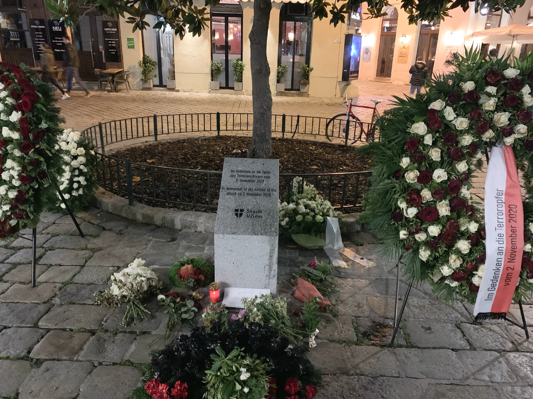 A street with a tree and a memorial stone commemorating the victims of the November 2020 attack.