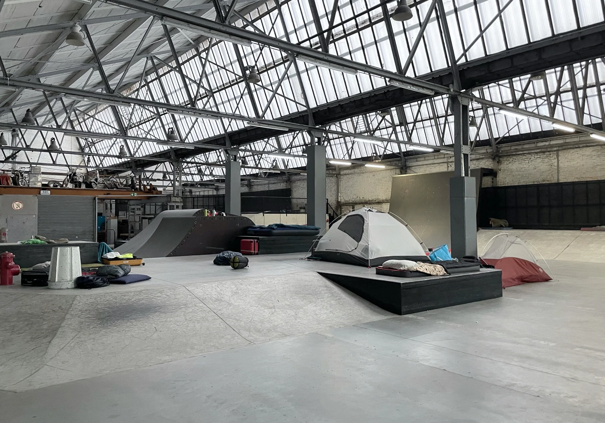 Foto of the sleeping area, a barren indoor skating and biking track with a few mattresses on the floor and the occasional tent set up.