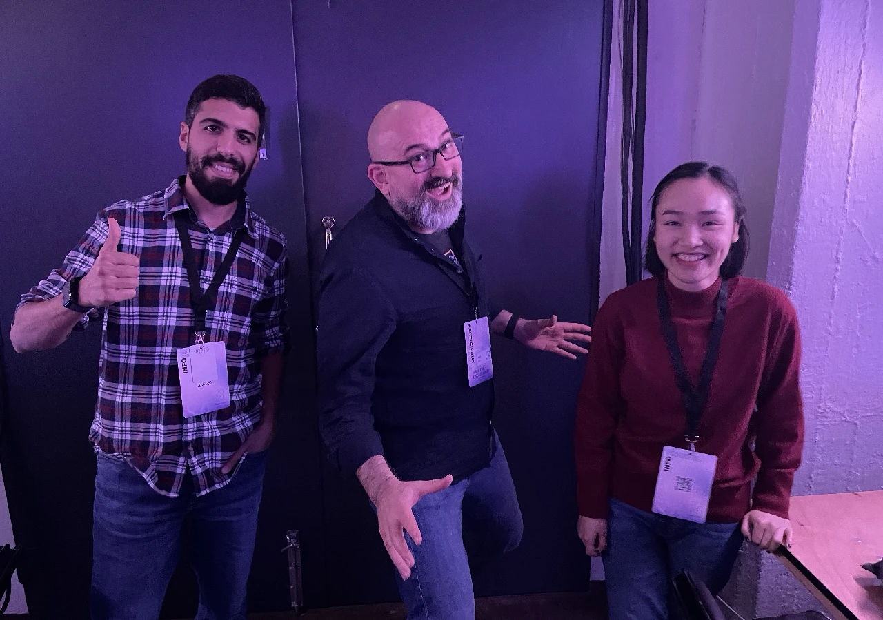A photo showing a young student with a black beard from Algeria, an older man with a gray beard from Austria, and a young lady from Vietnam. They are striking a pose for the camera.